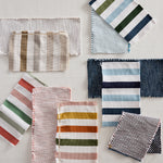 A flat-lay arrangement of multiple folded and striped fabric swatches in various colors and textures on a light off-white background.