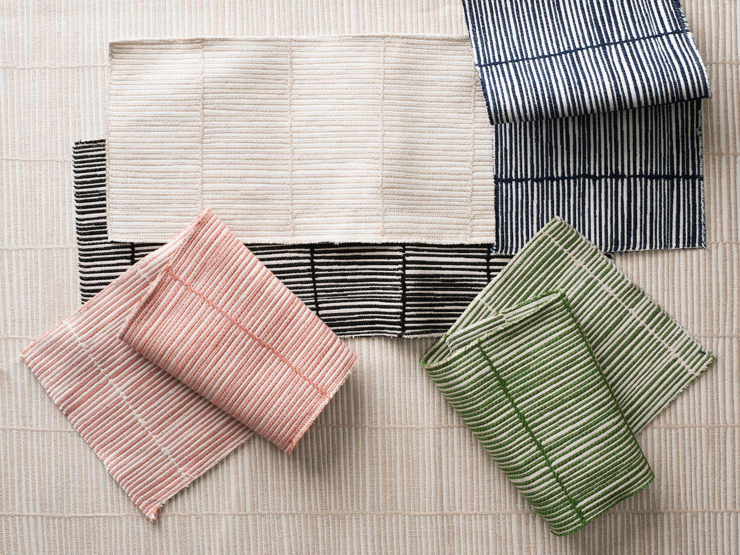 Styled flat lay with woven swatches of fabric with a fine bamboo stripe pattern in pink, green, black, and navy are arranged on beige background in the same pattern. 