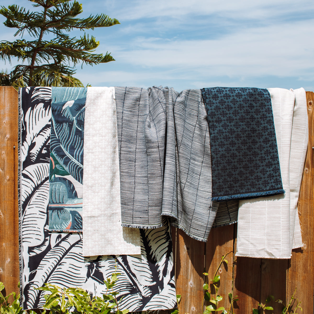 Several patterned in navy blue, teal and cream hanging outdoors on a wooden fence under a blue sky with clouds and green foliage in the foreground.