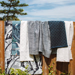 Several patterned in navy blue, teal and cream hanging outdoors on a wooden fence under a blue sky with clouds and green foliage in the foreground.