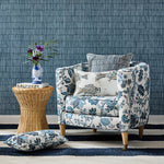 Detail of a living room scene, blue and white floral upholstered chair with patterned throw pillows, wicker side table with book and ceramic fish vase with flowers against blue bamboo striped wallpaper. 