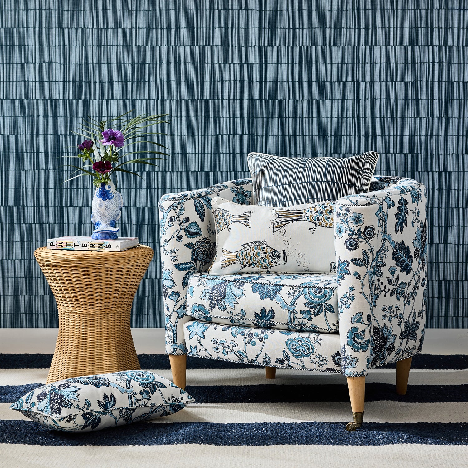 Detail of a living room scene, blue and white floral upholstered chair with patterned throw pillows, wicker side table with book and ceramic fish vase with flowers against blue bamboo striped wallpaper. 