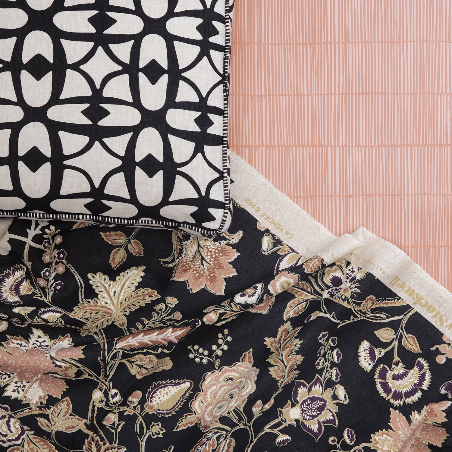 A close-up of floral and vine patterned fabric and decorative pillow with bold lattice design layered over a pink tonal striped wallpaper. 