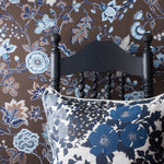 A close-up of a floral patterned pillow in front of a black wooden chair, set against brown wallpaper with blue and white floral designs.