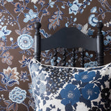 A close-up of a floral patterned pillow in front of a black wooden chair, set against brown wallpaper with blue and white floral designs.