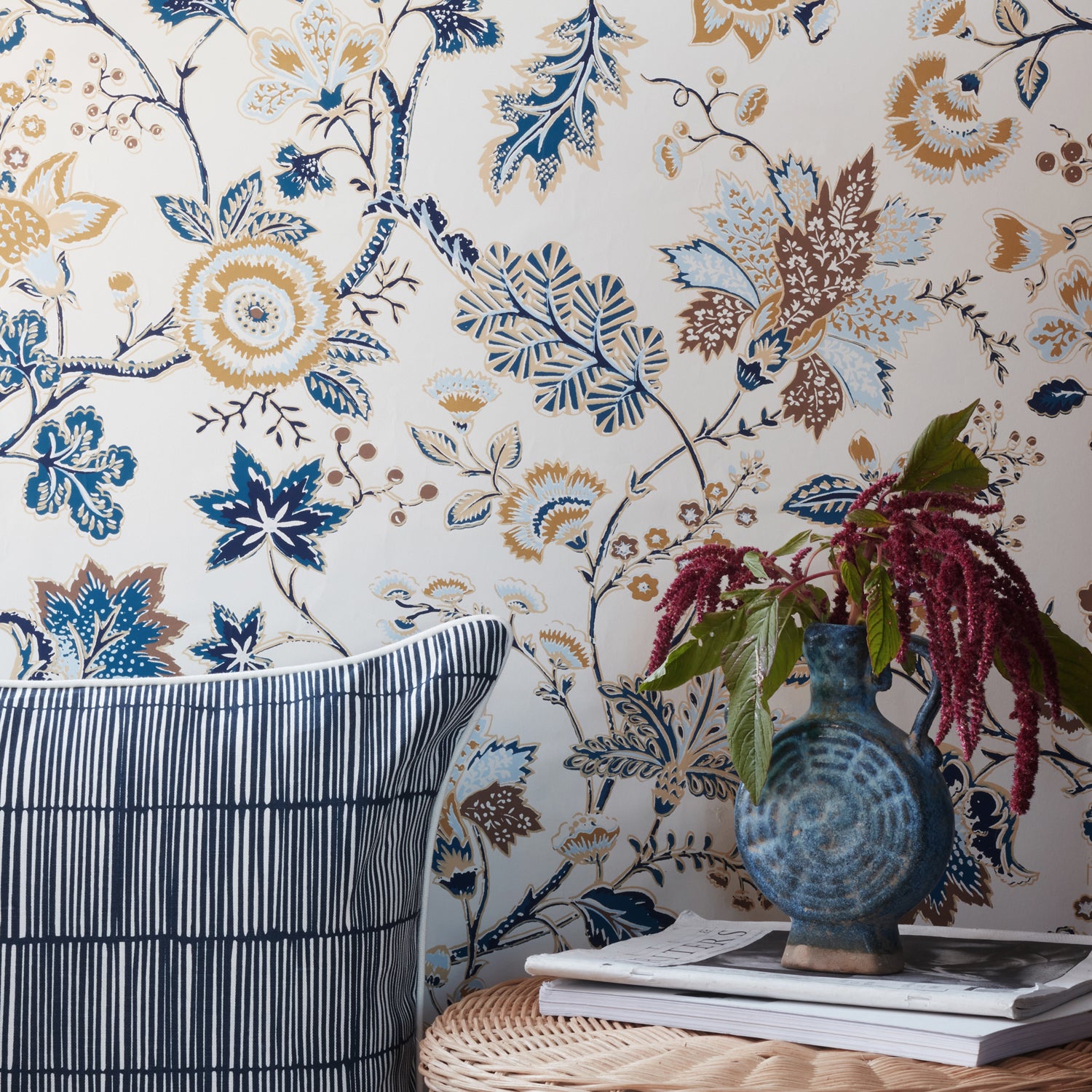 Floral wallpaper with blue, gold, and brown botanical patterns on a cream background, styled in a room setting with a striped pillow, a blue vase with burgundy flowers, and books on a woven table.
