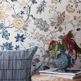 Floral wallpaper with blue, gold, and brown botanical patterns on a cream background, styled in a room setting with a striped pillow, a blue vase with burgundy flowers, and books on a woven table.