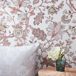 Floral wallpaper with a botanical pattern in dusty pink, terracotta, and beige on a white background, styled in a room setting with a patterned pillow and a small vase of flowers on a wicker side table.
