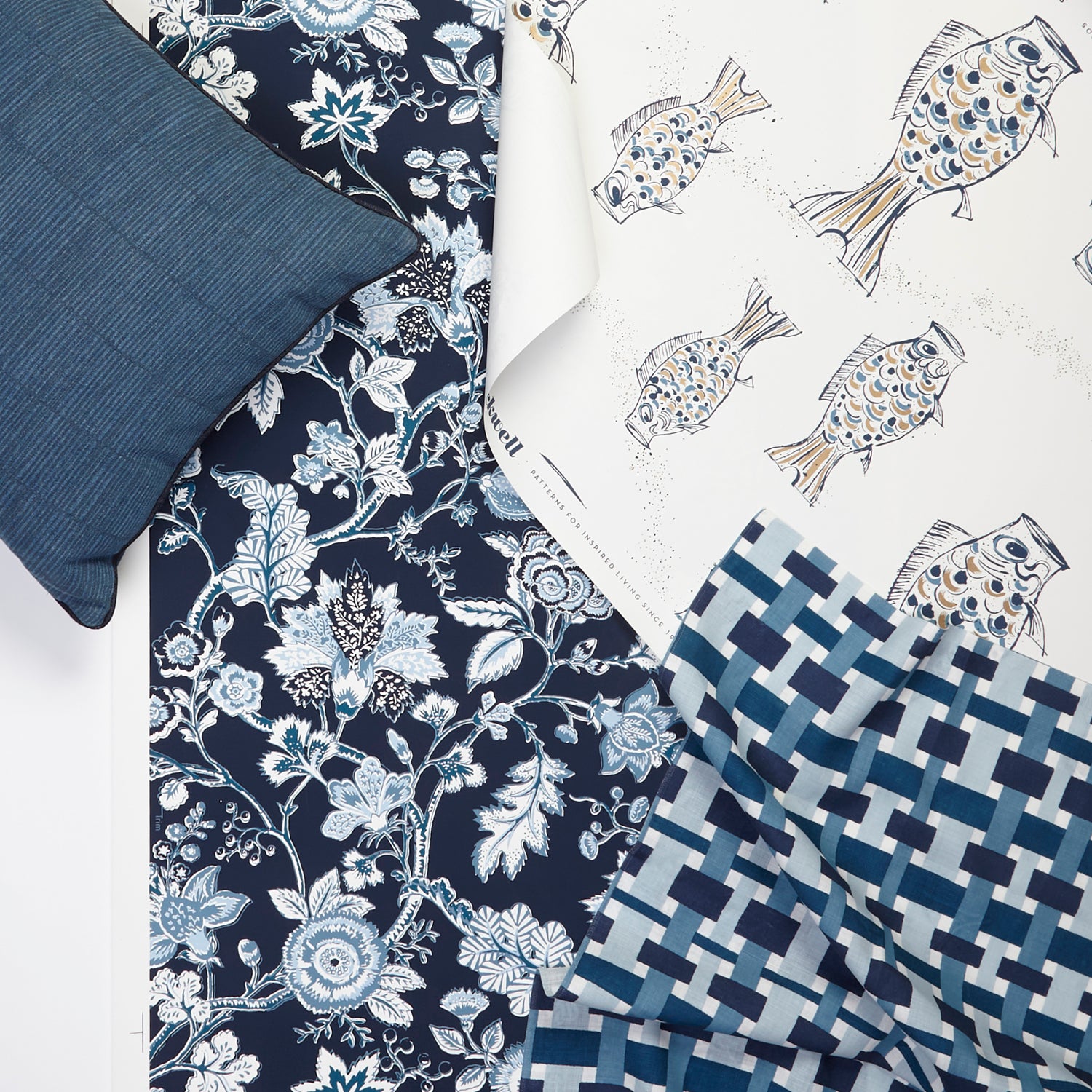 A styled flat-lay with basketweave plaid fabric layered with a playful fish print wallpaper and floral and vine wallpaper with a bamboo striped pillow in a palette of deep navy blues and white. 