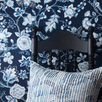 A close-up of a dark navy floral wallpaper with blue and white botanical patterns, styled behind a black wooden chair and a blue-and-white patterned throw pillow.