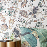 A styled interior scene showing a wall covered in botanical floral wallpaper with cream, green, and tan motifs, alongside a green banana-leaf-patterned cushion and a small side table with a drink and notebook.