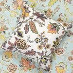 A square decorative throw pillow with a white base and a dense floral and botanical print in muted yellow, orange, green, and brown, set against a matching floral wallpaper in blue.