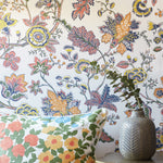 A close-up of a floral wallpaper with a vintage botanical pattern in orange, yellow, blue, and green on a white background, styled with a matching floral throw pillow and a textured gray vase.
