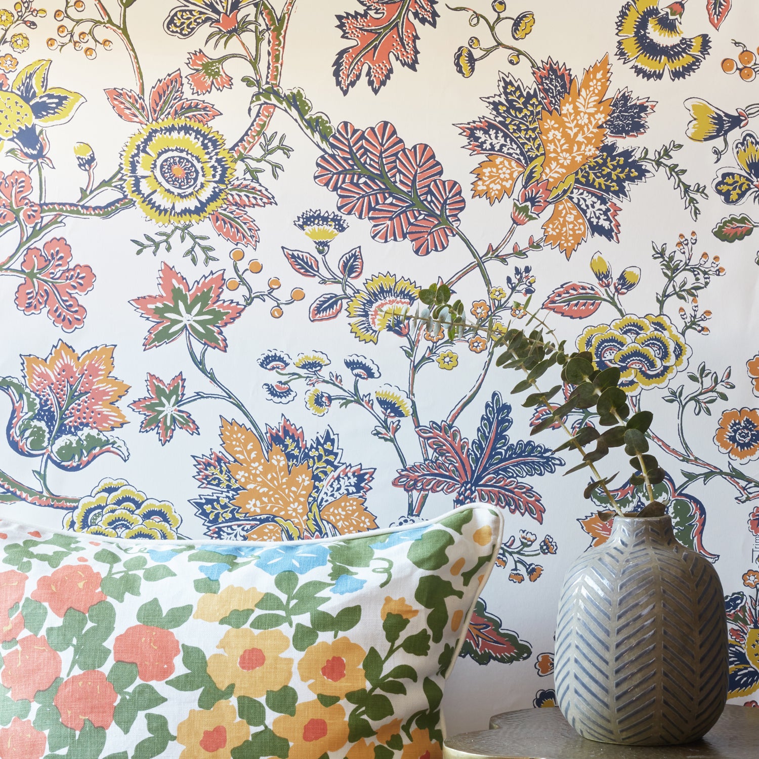 A close-up of a floral wallpaper with a vintage botanical pattern in orange, yellow, blue, and green on a white background, styled with a matching floral throw pillow and a textured gray vase.