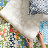 A styled flat lay showing a persons hands holding a wallpaper with layered throw pillows in a lattice print in shades of blue and beige, paired with a stack of brightly colored floral fabric, pillows and wallpapers. 