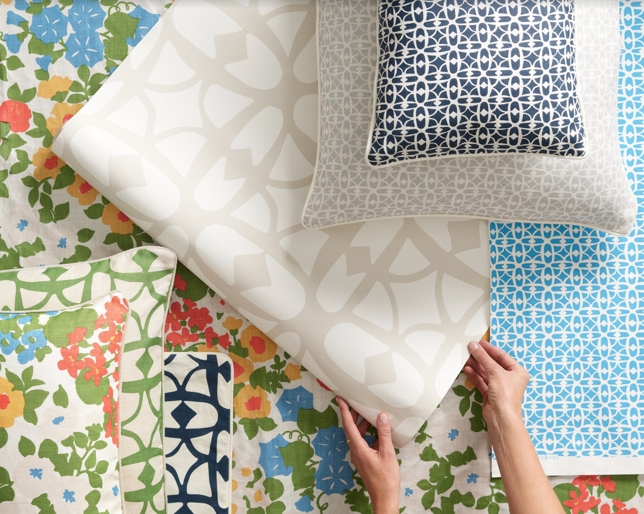 A styled flat lay showing a persons hands holding a wallpaper with layered throw pillows in a lattice print in shades of blue and beige, paired with a stack of brightly colored floral fabric, pillows and wallpapers. 