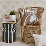 A styled interior scene with a tall woven rattan armchair holding a brown-and-white palm leaf throw pillow, next to a black-and-white striped side table with stacked books and a small plant. A second patterned pillow rests at the base of the chair against a beige patterned wallpaper background.