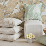 A styled interior scene with multiple decorative throw pillows in neutral and soft green tones, arranged around a cream upholstered cube against a tropical leaf wallpaper background. A small vase of white flowers sits on the floor.