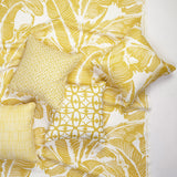A styled flat-lay of a yellow and white tropical leaf printed fabric with decorative throw pillows in mix of prints on a white background.