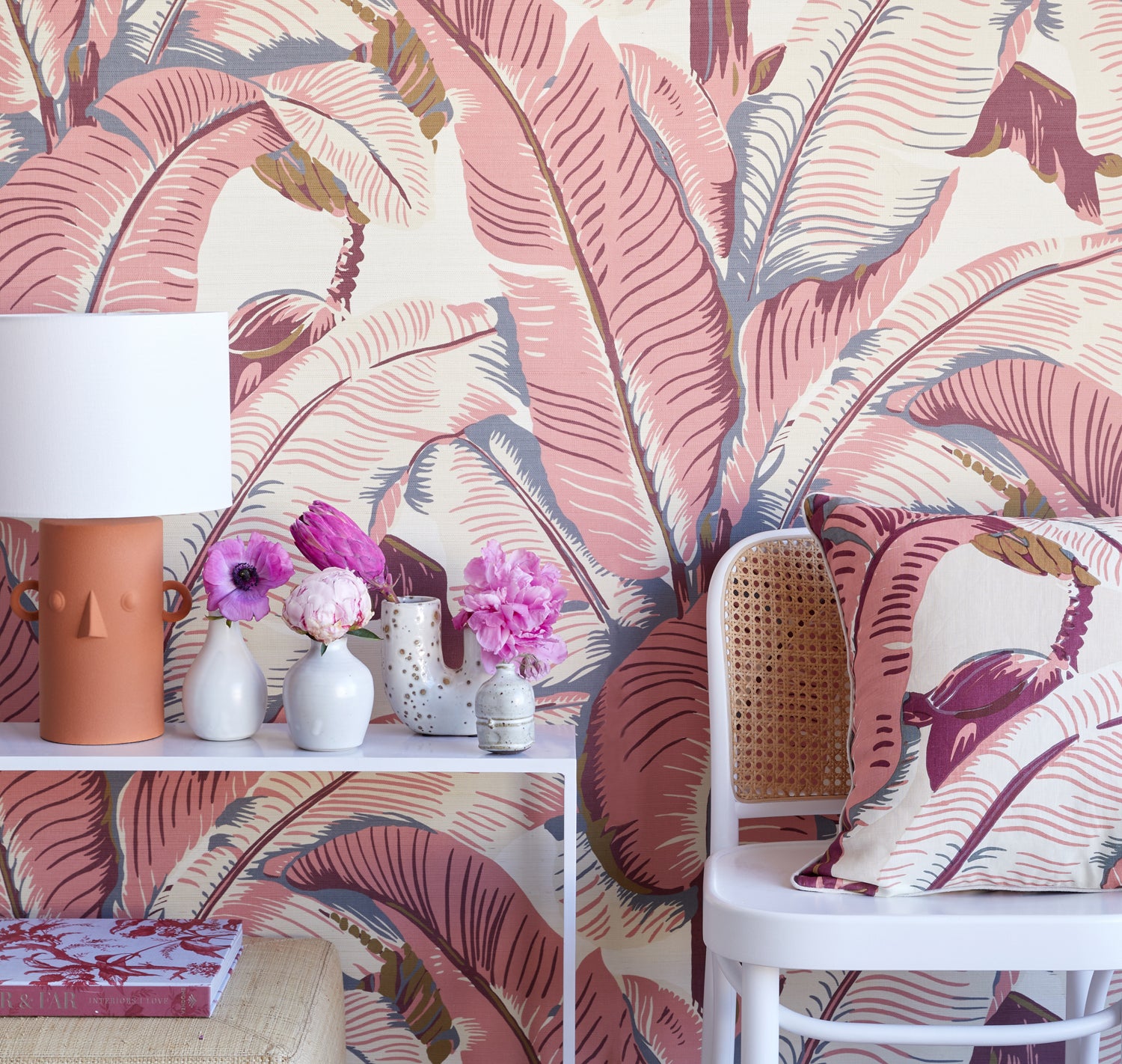 A styled interior scene featuring tropical wallpaper with large pink and mauve banana leaves on a cream background, a white side table with a terracotta lamp and small vases, and a white cane-back chair with a matching patterned pillow.