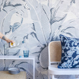 A styled interior scene with a large-scale tropical leaf wallpaper in gray and white, a navy floral throw pillow on a white cane-back chair, and a hand pouring water from a bottle into a glass on a white side table.