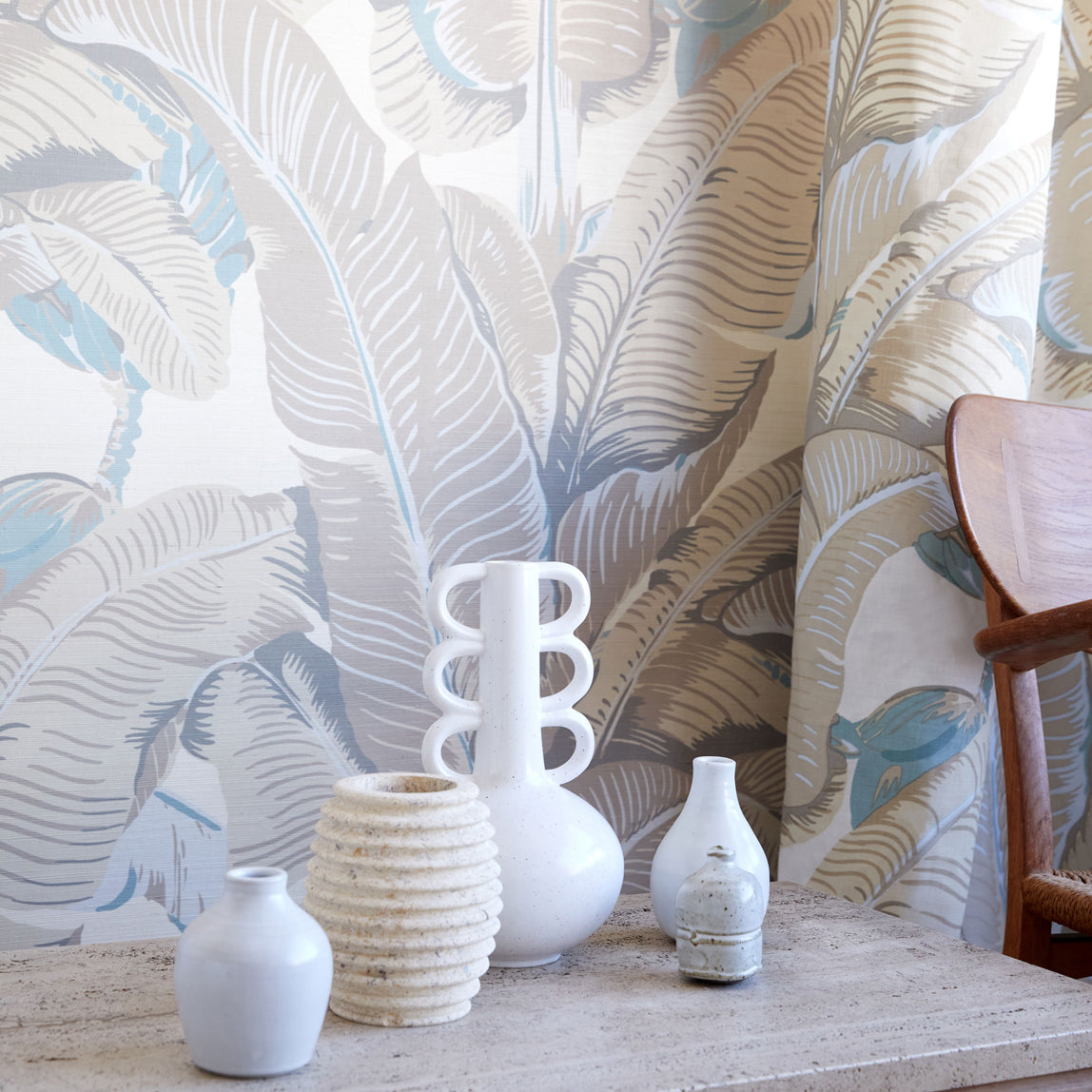 A styled interior scene showing a set of white ceramic vases and a textured beige vase arranged on a stone console table in front of a tropical leaf patterned wallpaper and curtain.