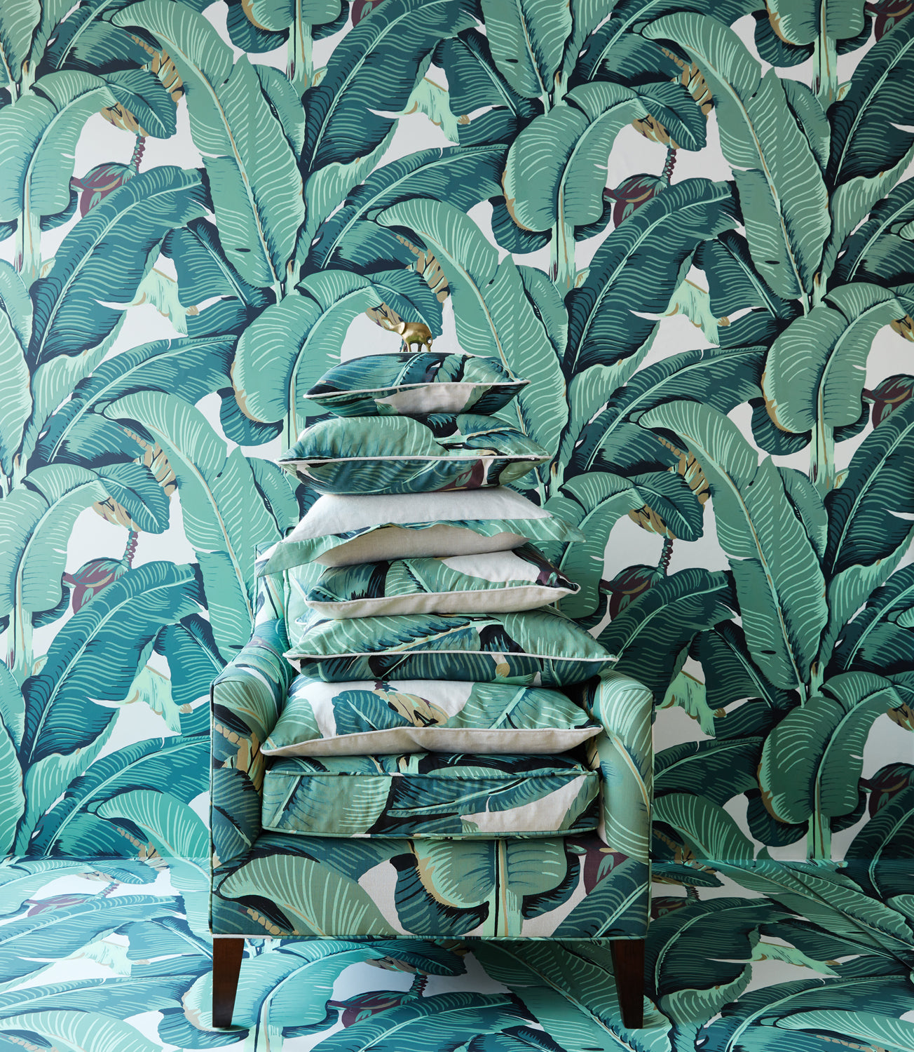 A stack of decorative throw pillows in a green and teal banana leaf print sits on a matching upholstered armchair with dark wood legs, set against a wall and floor covered in the same tropical leaf wallpaper.