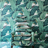 A stack of decorative throw pillows in a green and teal banana leaf print sits on a matching upholstered armchair with dark wood legs, set against a wall and floor covered in the same tropical leaf wallpaper.