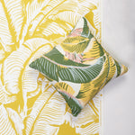 A square decorative throw pillow with a tropical banana leaf pattern in green, yellow, and white, styled on top of a matching yellow and white wallpaper.
