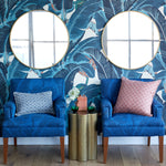 Two blue upholstered armchairs with patterned pillows are placed in front of a tropical leaf wallpaper wall with two round gold-framed mirrors. A small gold side table with green plants and a pink box sits between the chairs.