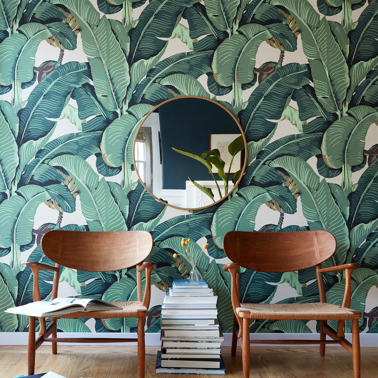 A styled interior scene featuring a bold green tropical banana leaf wallpaper on the wall, with two mid-century modern wooden chairs, a round gold-framed mirror, and a stack of books on a light wood floor.
