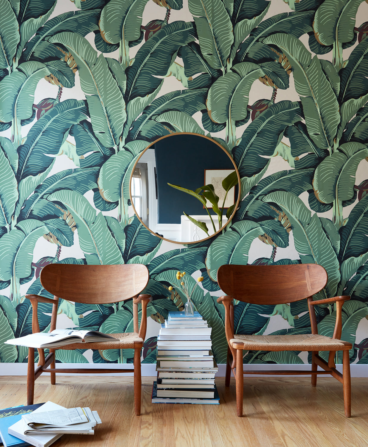 A styled interior scene featuring a bold green tropical banana leaf wallpaper on the wall, with two mid-century modern wooden chairs, a round gold-framed mirror, and a stack of books on a light wood floor.