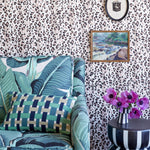A styled interior corner featuring a teal and green tropical leaf print armchair with a green plaid pillow, set against wallpaper with black animal spots. A small black-and-white striped side table with pink flowers is visible on the right, along with framed wall art.