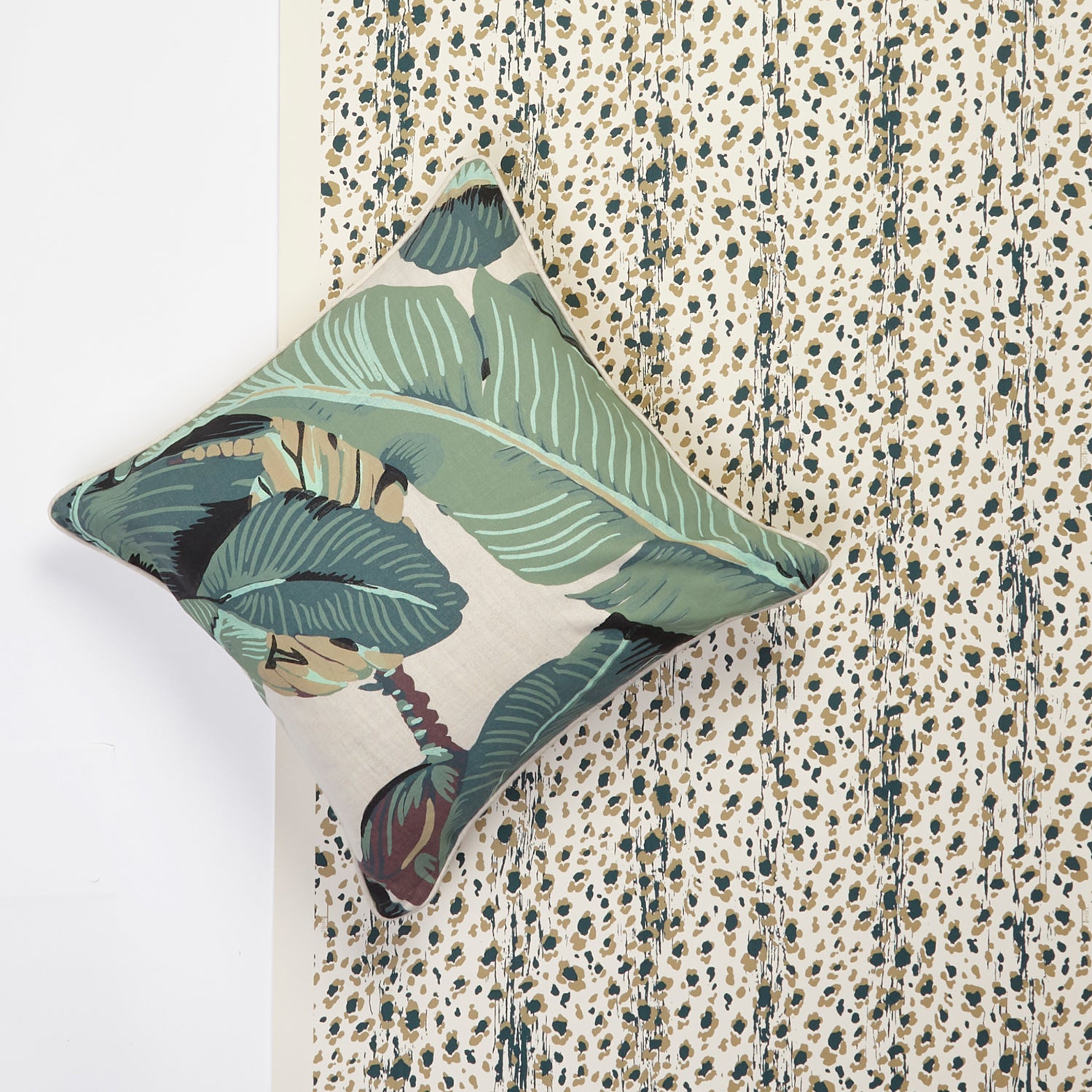 A square decorative throw pillow with a green tropical banana leaf pattern, styled on top of a tan and green leopard-print wallpaper. 