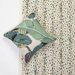 A square decorative throw pillow with a green tropical banana leaf pattern, styled on top of a tan and green leopard-print wallpaper. 