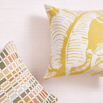 Two decorative throw pillows on a lwhite textured fabric. The larger pillow on the right features a bold yellow and white palm leaf pattern, while the smaller pillow on the left displays a geometric mosaic tile design in muted orange, beige, and olive tones.
