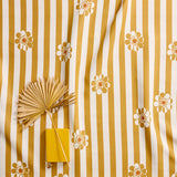 A styled flat-lay of a fabric with vertical mustard yellow and white stripes and scattered white flowers with orange centers. A yellow notebook and a dried palm leaf are placed on top as props.