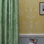 Green patterned curtain next to yellow wallpaper with a decorative vase on a table.