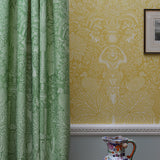 Green patterned curtain next to yellow wallpaper with a decorative vase on a table.