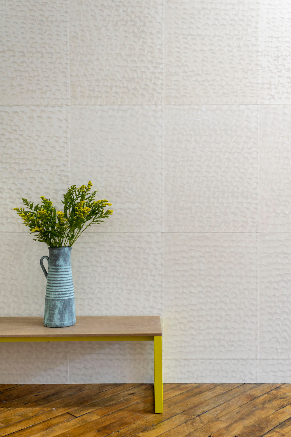 An end table with a vase of flowers stand in front of a wall papered in a repeating dappled texture in cream.
