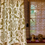 A curtain in a printed fabric in a lion, mixed animal and plant pattern in olive on a white field, against a window with with a reed shade and ceramic vases on the sill. 