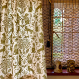 A curtain in a printed fabric in a lion, mixed animal and plant pattern in olive on a white field, against a window with with a reed shade and ceramic vases on the sill. 