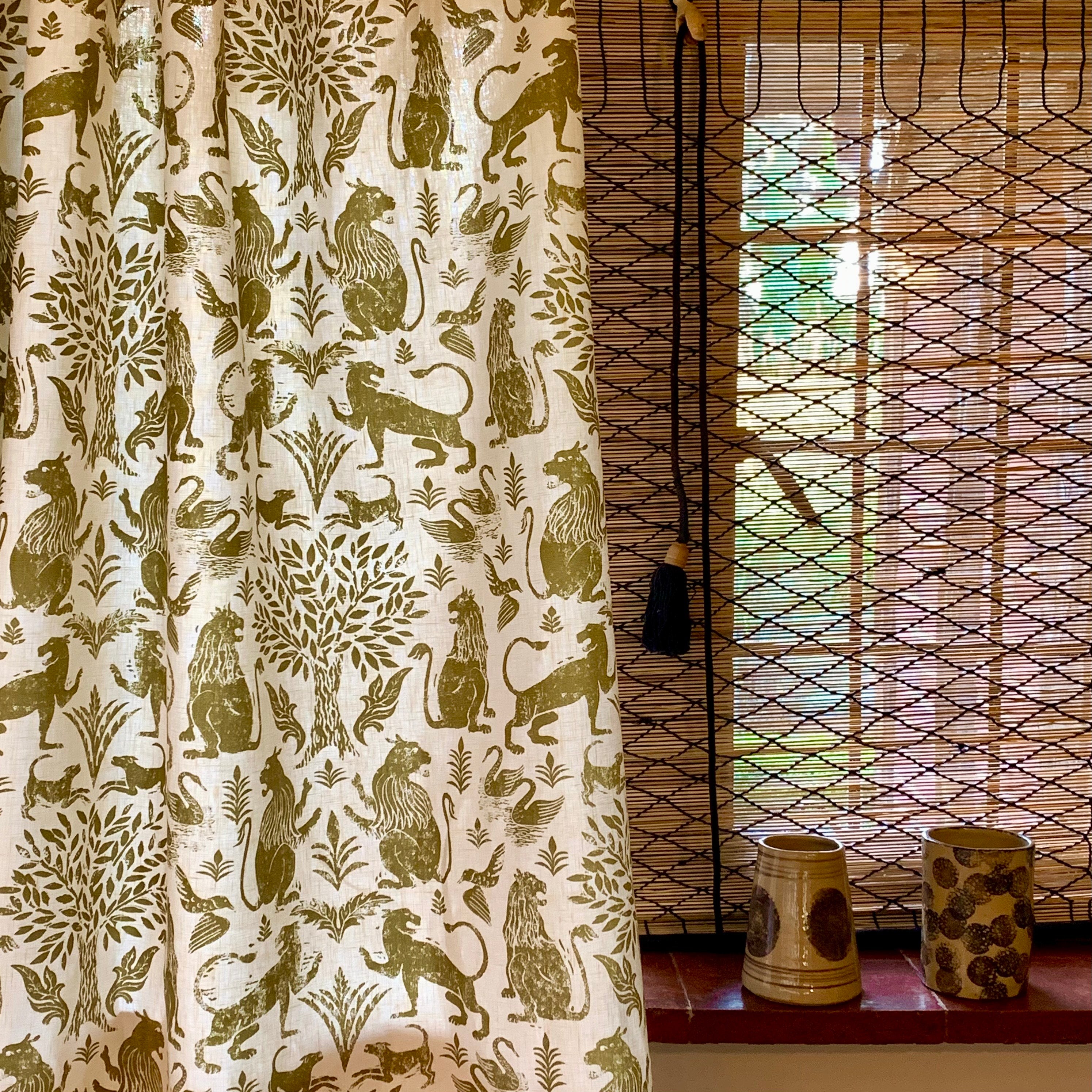 A curtain in a printed fabric in a lion, mixed animal and plant pattern in olive on a white field, against a window with with a reed shade and ceramic vases on the sill. 
