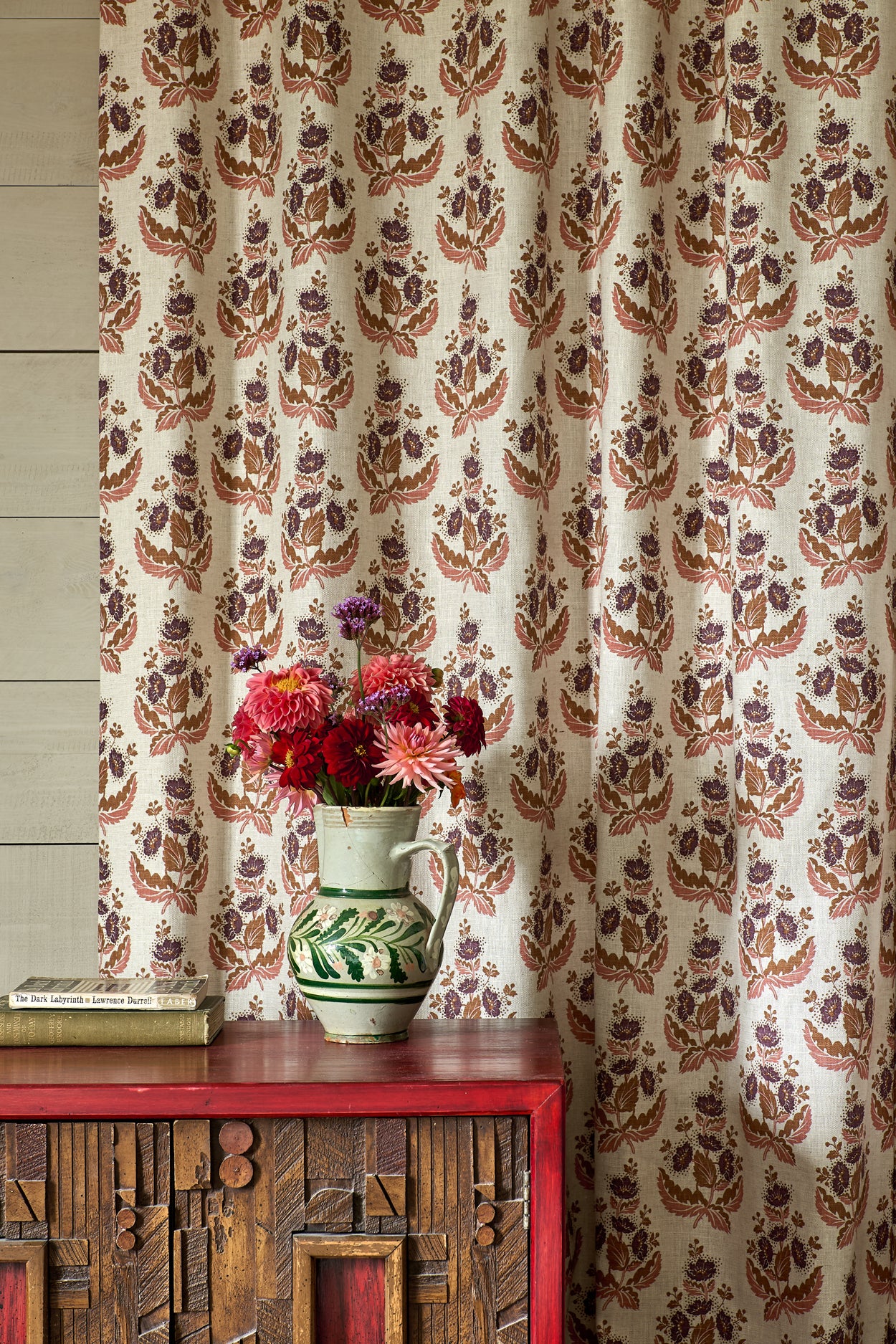 Decorative curtain with floral pattern in front of a wooden cabinet with a vase of flowers on top.