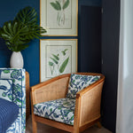 A wooden armchair with blue and white floral patterned cushions, set in a room with a dark blue wall, framed botanical prints, a potted monstera plant, and a black folding screen.
