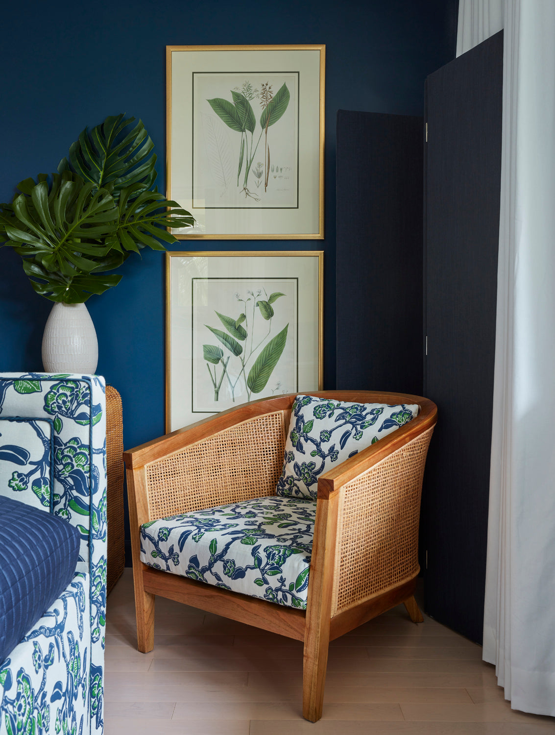 A wooden armchair with blue and white floral patterned cushions, set in a room with a dark blue wall, framed botanical prints, a potted monstera plant, and a black folding screen.