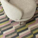 White bouclé side chair atop a handwoven rug with a stacked zig zag stripe in purple, lime green, cream and shades of grey. 