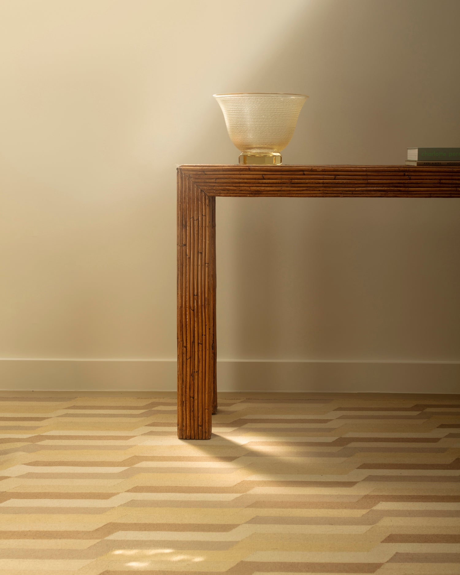 Bamboo console table against a white wall with glass vase and books atop a handwoven rug with a stacked zig zag stripe in shades of brown, taupe, and tan. 