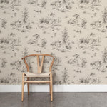 A wooden chair stands in front of a wall papered in a painterly shrub and tree print in charcoal on a cream field.
