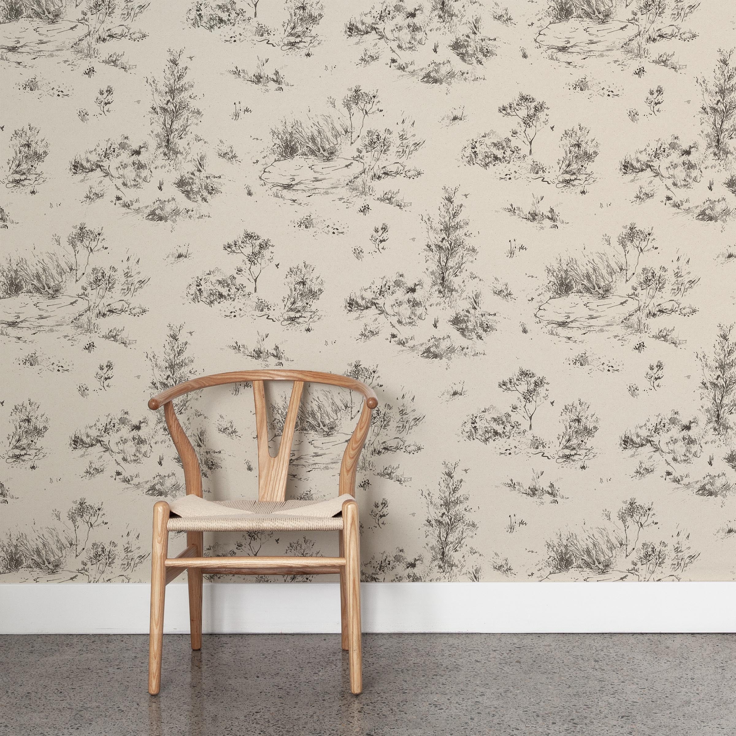 A wooden chair stands in front of a wall papered in a painterly shrub and tree print in charcoal on a cream field.